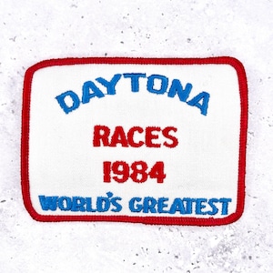 May include: A rectangular patch with a white background and a red border. The patch features the words "DAYTONA RACES 1984 WORLD'S GREATEST" in blue and red embroidered lettering. The patch is likely a vintage souvenir or collectible.