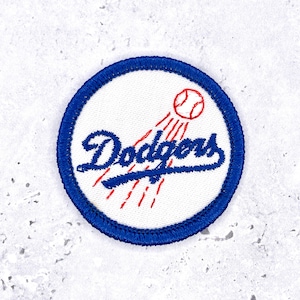 Può includere: Una toppa ricamata circolare con sfondo bianco e bordo blu. Il design presenta la parola Dodgers in corsivo blu, accompagnata da una pallina da baseball rossa ricamata e linee di movimento sulla superficie in tessuto bianco.