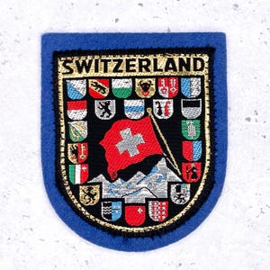 May include: Embroidered patch featuring the word "SWITZERLAND" above a shield design. The shield displays the Swiss flag, mountains, and various emblems. The patch has a blue border and gold trim.