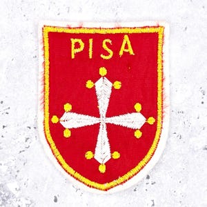 May include: A shield-shaped embroidered patch with a red background and a yellow border. The text PISA is stitched in yellow at the top. Centered is a white cross with flared arms, each tipped with a small yellow circle at the ends.