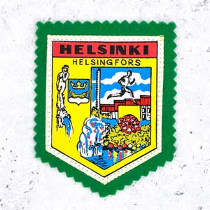 May include: A green felt patch with a shield-shaped design featuring the text HELSINKI and HELSINGFORS. The graphic includes a yellow background, a blue shield with a gold crown and boat, a running figure, a fountain, and a red floral element.