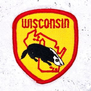 Può includere: Toppa ricamata con la scritta "WISCONSIN" in rosso, arcuata su sfondo giallo. Sono presenti anche un contorno rosso dello stato e un tasso bianco e nero. La toppa ha un bordo rosso.