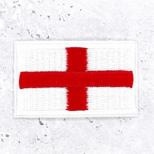 May include: A rectangular embroidered patch featuring the flag of England, which consists of a bold red cross centered on a white background. The stitching is dense and uniform, creating a textured surface with clearly defined edges.