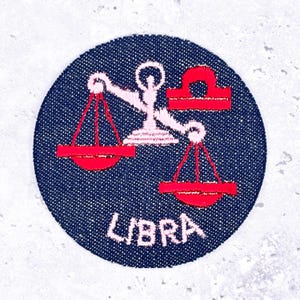May include: A round, navy blue embroidered patch featuring the Libra zodiac symbol. The design includes a pink scale and the word "LIBRA" in pink lettering. The patch is circular and has a textured appearance.