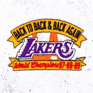 May include: Embroidered patch featuring the word "Lakers" in purple and white, with a yellow banner above that reads "Back to Back & Back Again." A red banner below says "World Champions 87-88-89."