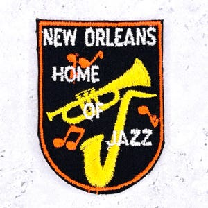 May include: Embroidered patch with the text "NEW ORLEANS HOME OF JAZZ". The patch features a yellow saxophone and trumpet, with orange musical notes on a black background, framed by an orange border.