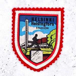 May include: A shield-shaped embroidered patch with a red scalloped border. The center features a graphic of the Helsinki Stadion with a tall tower, a statue of a runner on a pedestal, and green trees. The text reads HELSINKI Helsingfors STADION.