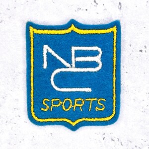 Könnte beinhalten: Blauer Filzaufnäher mit gelbem Rand und weißer gestickter Schrift. Der Aufnäher zeigt die Buchstaben "NB" über dem Wort "SPORTS". Der Aufnäher hat eine Schildform mit dekorativer Ober- und Unterseite.