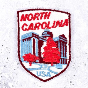 May include: Embroidered patch with the words "NORTH CAROLINA" in red, above a building and trees design. The patch has a white background with blue and maroon detailing, and the word "USA" is at the bottom.
