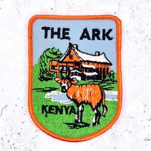 Può includere: Una toppa ricamata rettangolare con angoli arrotondati e bordo arancione. Raffigura un cielo azzurro, un edificio in legno marrone, erba verde e un'antilope bongo marrone e bianca. Il testo riporta THE ARK e KENYA in nero.