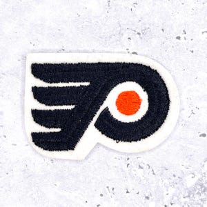 May include: Embroidered patch featuring the Philadelphia Flyers logo. The logo is a stylized black 'P' and wing design with an orange circle in the center, outlined in white. The patch is on a white background.