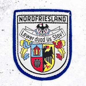 May include: A rectangular embroidered patch with a blue border featuring the text NORDFRIESLAND at the top. Below, a banner reads Lewer duad üs Slav! above a shield divided into sections with a crown, a black eagle, and floral motifs.