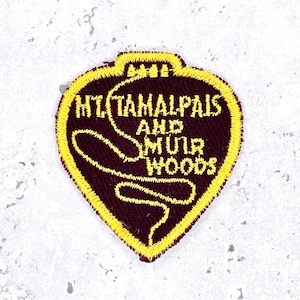 May include: A shield-shaped embroidered patch with a dark maroon background and a bright yellow border. The text "MT. TAMALPAIS AND MUIR WOODS" is stitched in yellow thread, accompanied by a yellow embroidered line that curves across the surface.