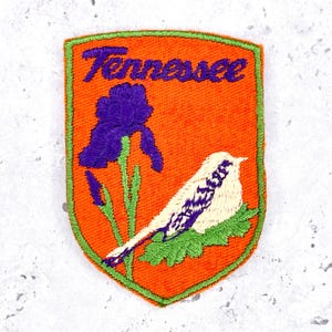 May include: A shield-shaped embroidered patch with an orange background and a bright green border. It features the word Tennessee in purple script at the top, a purple iris flower on a green stem, and a white bird with purple markings on a green leaf.