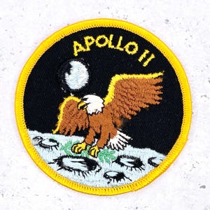 Pode incluir: Remendo redondo com fundo preto e borda amarela. Apresenta uma águia com asas marrons e cabeça branca, pousando na lua. O texto "APOLLO II" é amarelo.