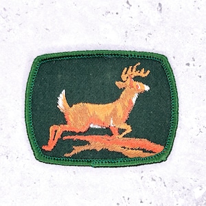 May include: Embroidered patch depicting a leaping deer in orange and white, set against a dark green background. The deer is jumping over a log, with a green border. Suitable for jackets, bags, or other fabric items.