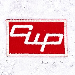 May include: A rectangular embroidered patch featuring a vibrant red background with a white stitched border. The word "Cup" is displayed in the centre in a bold, stylised white font with thick, clean embroidery lines throughout the design.