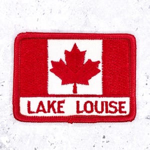 May include: A rectangular embroidered patch featuring a red and white design inspired by the Canadian flag with a central red maple leaf. The lower white section contains the red embroidered text LAKE LOUISE in a bold, sans-serif font.