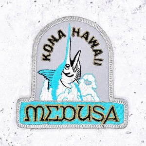 Op de afbeelding: Geborduurde patch met een marlijn die uit de oceaan springt. De patch heeft een lichtgrijze achtergrond met de woorden "KONA HAWAII" in gouden letters. De onderkant van de patch leest "MEDUSA" in gouden letters op een turquoise achtergrond.
