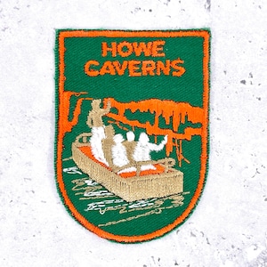 Pode incluir: Patch bordado com as palavras "HOWE CAVERNS" em laranja sobre um fundo verde. O patch apresenta um barco com passageiros e formações de cavernas. O patch tem uma borda laranja.