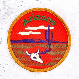 Può includere: Una toppa ricamata circolare con uno sfondo rosso brillante e la parola Arizona cucita in verde nella parte superiore. Il design include un teschio bianco, un cactus saguaro viola, una catena montuosa viola e due nuvole bianche.