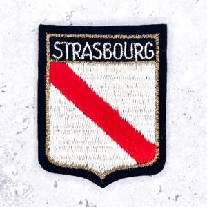 May include: A shield-shaped embroidered patch featuring a black border with the word STRASBOURG stitched in white at the top. The center displays a white background with a diagonal red stripe, all outlined by a thin metallic gold thread border.