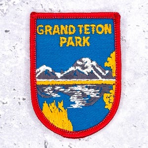 Puede incluir: Parche bordado con fondo azul con el texto amarillo "GRAND TETON PARK". El parche representa una cordillera reflejada en un lago, con detalles amarillos y blancos, enmarcado por un borde rojo.