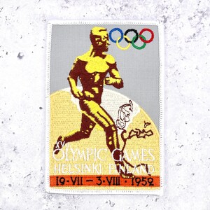 May include: A rectangular embroidered patch featuring a golden runner silhouette against a light gray background. It displays the Olympic rings, the text XV OLYMPIC GAMES HELSINKI FINLAND, and the dates 19-VII-3-VIII-1952 in a dark orange band.