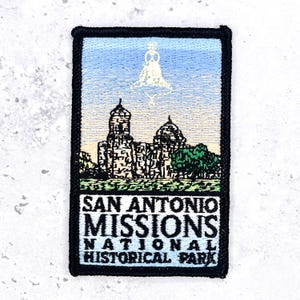 Op de afbeelding: Een rechthoekige geborduurde patch met een zwarte rand die het San Antonio Missions National Historical Park afbeeldt. Het ontwerp bevat een stenen missiegebouw, een klok en groene bomen onder een blauwe lucht met tekst eronder.