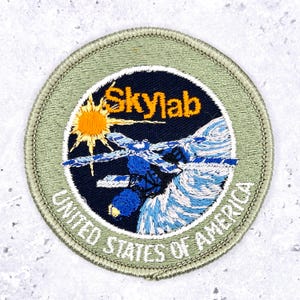 May include: A circular embroidered patch featuring a dark blue center with a yellow sun, a blue and white space station, and the text Skylab. The border is light olive green with the white text United States of America embroidered along the bottom.
