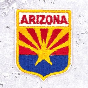 May include: Embroidered Arizona state flag patch. The patch features the word "ARIZONA" in red on a white background. Below, a red and yellow sunburst design is above a blue base with a yellow star.