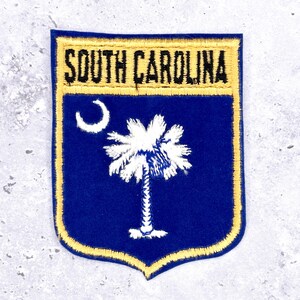 May include: A blue and gold embroidered patch featuring the words "SOUTH CAROLINA" at the top. The patch has a shield shape and includes a white crescent moon and a white palmetto tree.