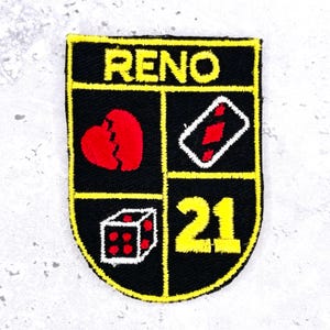 May include: A black embroidered patch with a yellow border and the text RENO at the top. The design is divided into three sections featuring a red heart, a white playing card with a red diamond, a white die with red dots, and the yellow number 21.