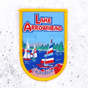 Puede incluir: Un parche azul con las palabras "LAKE ARROWHEAD" en rojo y blanco. El parche presenta veleros en un lago, árboles verdes y la palabra "CALIFORNIA" en la parte inferior. El parche tiene un borde amarillo.