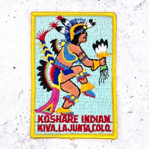May include: Embroidered patch featuring a Native American figure in colorful ceremonial attire. The figure is depicted in motion, holding a fan, with text that reads "Koshare Indian Kiva La Junta Colo" in red letters. The patch has a light blue background and a yellow border.