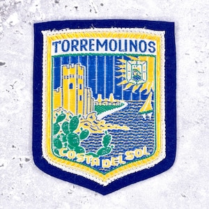 May include: Embroidered patch with a navy blue border. The patch features the text "TORREMOLINOS" at the top and "COSTA DEL SOL" at the bottom. The design includes a yellow castle, blue sea, green cacti, and a sailboat.