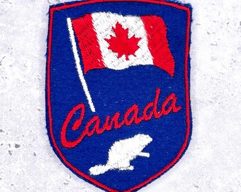 May include: A blue, shield-shaped patch with a red border. The patch features a Canadian flag, the word "Canada" in red script, and a white beaver. The flag has a red maple leaf on a white background.