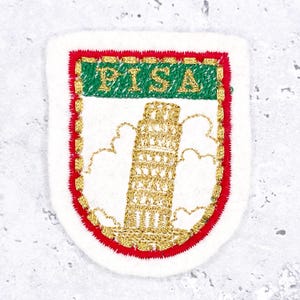 Könnte beinhalten: Gestickter Aufnäher mit dem Schiefen Turm von Pisa. Der Aufnäher hat einen weißen Hintergrund mit rotem Rand. Das Wort "PISA" ist goldfarben auf einem grünen Banner gestickt. Der Turm ist mit goldenem Faden detailliert.