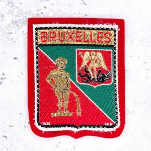 Op de afbeelding: Een rechthoekige geborduurde patch met een rode rand en de tekst BRUXELLES in gouddraad. Het ontwerp is diagonaal verdeeld in rood en groen en toont een gouden figuur van Manneken Pis en een gouden embleem van de Heilige Michaël.