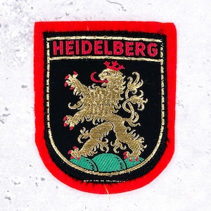 May include: A rectangular embroidered patch with a red border featuring a black shield-shaped centre. The text HEIDELBERG is stitched in red at the top. A gold lion with a red tongue and claws stands on a green mound within the shield.