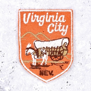 以下が含まれることがあります： オレンジ色の刺繍パッチ。白い筆記体で「Virginia City」と書かれています。馬に引かれた幌馬車が描かれており、下部には「NEV」の略語があります。白い縁取りがあります。