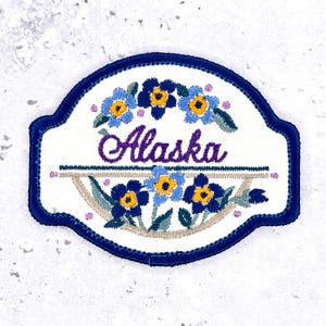 May include: An embroidered patch with a dark blue border on a white background. The center features the word Alaska in purple script, surrounded by blue and yellow flowers with green leaves. A horizontal line separates the floral designs.