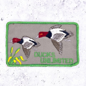 May include: Embroidered patch showing two ducks in flight, with red heads, white and grey wings, and brown bodies. The patch has a grey background, green trim, and the words "DUCKS UNLIMITED" in green.