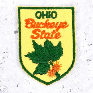 May include: A shield-shaped embroidered patch with a yellow background and a dark green border. The text OHIO is stitched in dark green, while Buckeye State is in orange. A detailed dark green buckeye leaf and two orange spiky fruits are below.