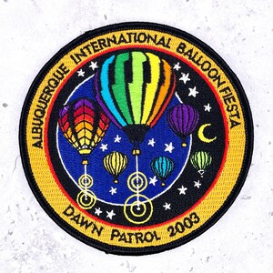 May include: A circular embroidered patch featuring the text Albuquerque International Balloon Fiesta and Dawn Patrol 2003. The design displays multiple colorful hot air balloons against a dark blue starry night sky with a yellow crescent moon.