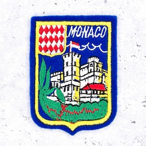 Op de afbeelding: Een Monaco-patch met een blauwe rand en een gele omtrek. De patch heeft een rood-wit geblokt ontwerp, het woord "MONACO", een kasteel en een vlag. Groen gebladerte staat onderaan.