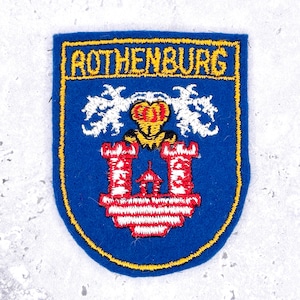 May include: A blue embroidered patch featuring the text ROTHENBURG in yellow at the top. The design includes a yellow crown above a red and white castle structure with two towers, all outlined in yellow thread on a blue felt background.