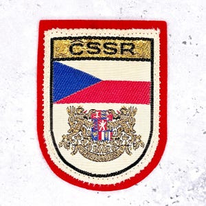 Könnte beinhalten: Ein rechteckiger Aufnäher mit rotem Rand und einer weißen, goldenen und schwarzen Perlenborte. Der Aufnäher zeigt die tschechische Flagge und den Text "CSSR" in Gold. Unter der Flagge befindet sich ein detailliertes Emblem.