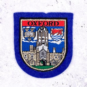 May include: A blue felt patch with a gold border and the word "OXFORD" in red. The patch features a detailed illustration of a building and crests, likely a university emblem. The patch is a shield shape.