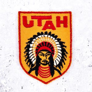 Pode incluir: Patch bordado com a palavra "UTAH" em vermelho sobre uma ilustração de um chefe nativo americano. O patch tem um fundo amarelo e uma borda vermelha. O design apresenta um cocar.